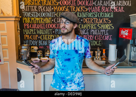 Giovane maschio server dietro il contatore alla gelateria con due vuota del gelato scoop nelle sue mani in un cappello e cravatta tinto camicia. Foto Stock