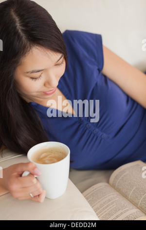 Giovane donna asiatica sdraiato sul divano a leggere un libro mentre tiene il suo caffè Foto Stock