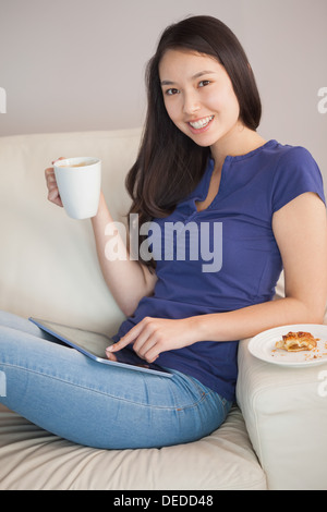 Felice giovane donna asiatica utilizzando il suo tablet pc e tenendo la tazza di caffè Foto Stock