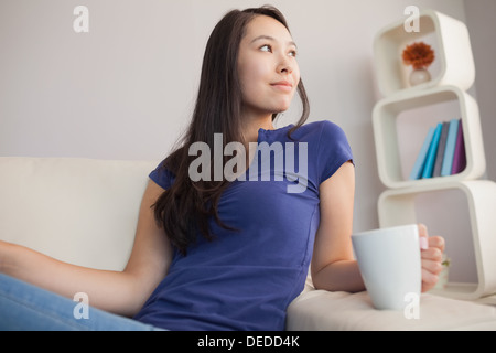 Attraente giovane donna asiatica seduta sul divano holding mug Foto Stock