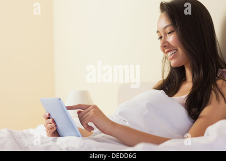 Sorridente giovane donna asiatica seduta nel letto utilizzando un tablet pc Foto Stock