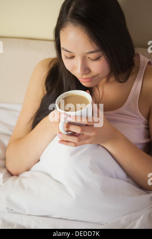 Allegro giovane donna asiatica seduta nel letto annusando il suo caffè del mattino Foto Stock