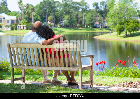 Vista posteriore di una felice romantico senior americano africano giovane seduto su una panchina nel parco che abbraccia guardando un lago Foto Stock