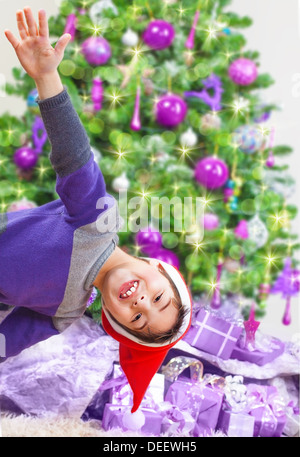 Felice ragazzo vicino ad albero di Natale, indossando red Santa hat, festeggiare il nuovo anno a casa, molti diversi doni, vacanze invernali concept Foto Stock