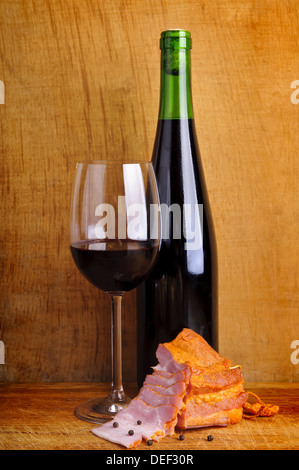 Ancora vita composizione con vetro e una bottiglia di vino rosso e prosciutto affumicato su un vintage backround in legno Foto Stock