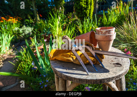 Attrezzi per giardinaggio utensili, guanti e vasi di fiori appoggiati su uno sgabello in un giardino verde Foto Stock