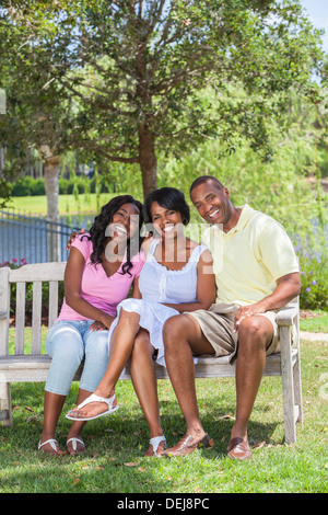 Un felice Africano nero americano della famiglia di due genitori e una ragazza seduta del bambino assieme al di fuori su una panchina nel parco di ridere Foto Stock