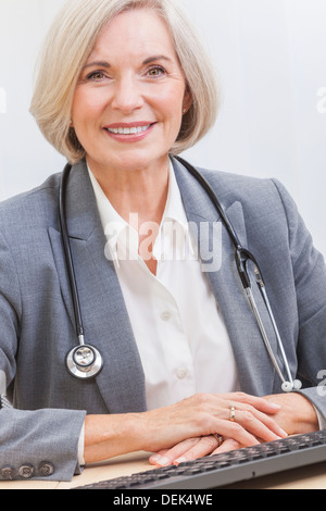 Un senior medico donna seduti ad una scrivania in un ufficio con un computer, indossa una tuta e stetoscopio Foto Stock