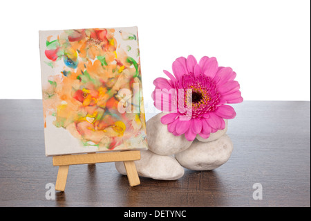 Pila di rocce bianche su un marrone scuro scaffale di legno decorato con una pink gerbera e un dipinto su un cavalletto Foto Stock