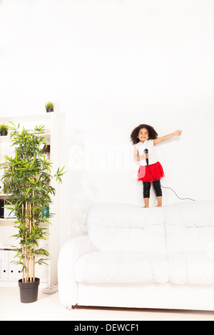 Carino piccolo Africano nero tre anni ragazza con microfono di eseguire in piedi sul divano con mano sollevata Foto Stock