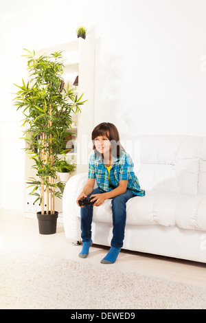 Felice 6 anni vecchio ragazzo di giocare con i videogiochi tenendo il controller di gioco seduti sul divano bianco in salotto Foto Stock