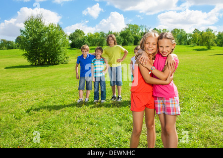 Due lieti di nove anni le ragazze in piedi nel parco avvolgente con un gruppo di ragazzi amici in piedi sullo sfondo nel parco sulla giornata di sole Foto Stock