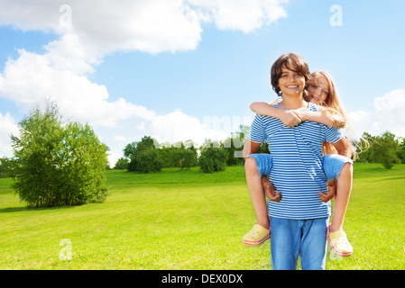 Scuola di giovane età i bambini ragazza ride su la parte posteriore del ragazzo e abbraccio guy, felice in piedi al di fuori del parco sulla soleggiata giornata estiva Foto Stock