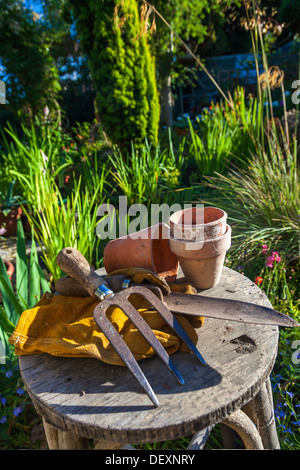 Attrezzi per giardinaggio utensili, guanti e vasi di fiori appoggiati su uno sgabello in giardino verde con una serra al di fuori della messa a fuoco in background Foto Stock