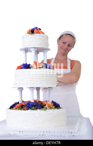 Baker lady dando ad una torta di nozze ultimo aspetto fiero nel suo grembiule bianco e bandana, torta ha fondente volant sul lato e Foto Stock