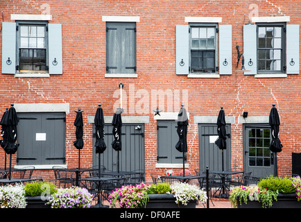 Un vecchio edificio in mattoni grigi con porte e persiane e chiuso nero ombrelloni dalle tabelle sul patio con fiori in scatole della piantatrice Foto Stock