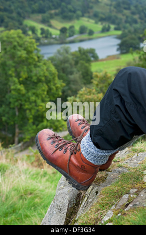 Primo piano di persona uomo camminatore indossando stivali da camminata seduto su un bordo di scogliera caduto nel Lake District National Park Inghilterra Regno Unito Gran Bretagna Foto Stock