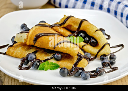 Due frittelle con mirtilli, menta e glassa di cioccolato su una piastra bianca, tovagliolo, vaso sullo sfondo delle tavole di legno Foto Stock