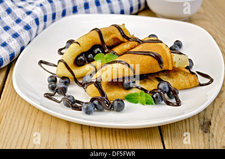 Due frittelle con mirtilli, menta, glassa di cioccolato su una piastra bianca, tovagliolo, vaso sullo sfondo delle tavole di legno Foto Stock