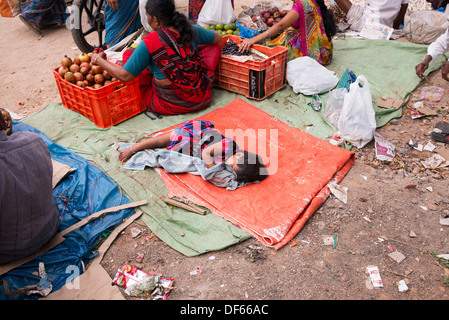 Giovane ragazza indiana in un villaggio di mercato addormentato sul pavimento mentre madre vende frutta . Puttaparthi, Andhra Pradesh, India Foto Stock