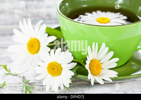 Primo piano di una tazza con erbe tè camomilla servita nel verde della tazza di ceramica e alcuni fiori selvatici sul dipinto di bianco di superficie in legno Foto Stock