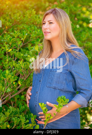 Ritratto di attraente donna incinta nel parco, femmina in attesa di toccare pancia sul fogliame verde, sfondo il concetto di amore Foto Stock