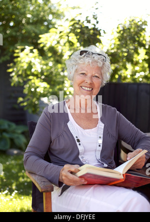 Donna anziana relax su una sedia in giardino con un libro guardando la telecamera sorridendo - Senior donna lettura all'aperto Foto Stock