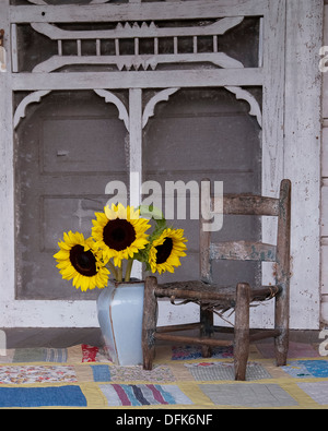 Una vecchia sedia, Vaso con girasoli, una trapunta su un portico di ruggine con una porta dello schermo come sfondo. Foto Stock