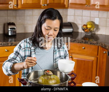 Foto di donna matura la cottura di melone invernale dal recipiente per la cottura a vapore con cucchiaio grande in mano e cucina di quercia rossa armadi in background Foto Stock