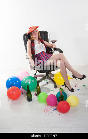 Party goer indossando un kilt e bere vino rosso da un grande bicchiere seduto in una sedia da ufficio Foto Stock