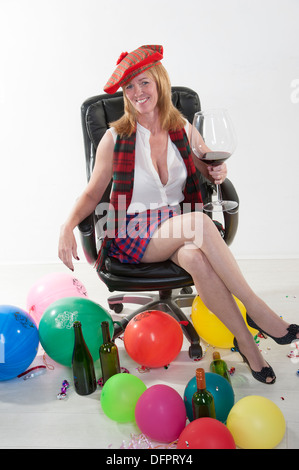 Party goer indossando un kilt e bere vino rosso da un grande bicchiere seduto in una sedia da ufficio Foto Stock