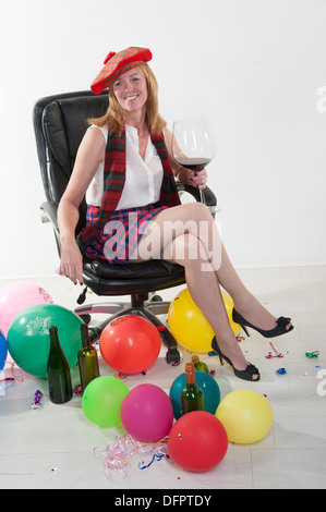 Party goer indossando un kilt e bere vino rosso da un grande bicchiere seduto in una sedia da ufficio Foto Stock