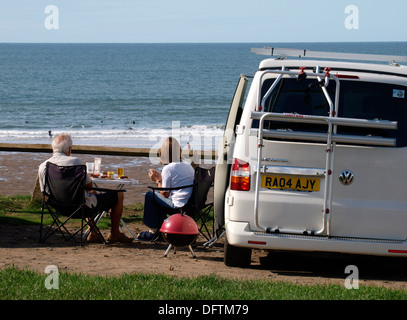 Coppia matura avente un barbecue accanto a loro VW Transporter van, Widemouth Bay, Bude Cornwall, Regno Unito Foto Stock