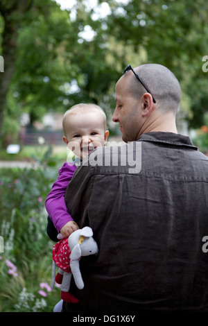 Baby girl ridendo e aggraffatura giocattolo morbido, guardando oltre il padre della spalla. Foto Stock