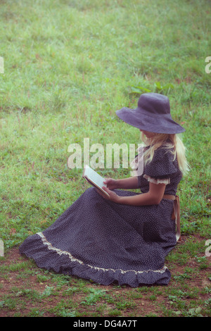 Una donna in un romantico abito è seduto su un prato, la lettura di un libro Foto Stock