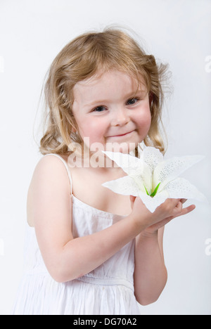 Bella innocente bambina vestita di bianco tenendo un grande fiore alla sua guancia, guardando direttamente alla fotocamera, REGNO UNITO Foto Stock