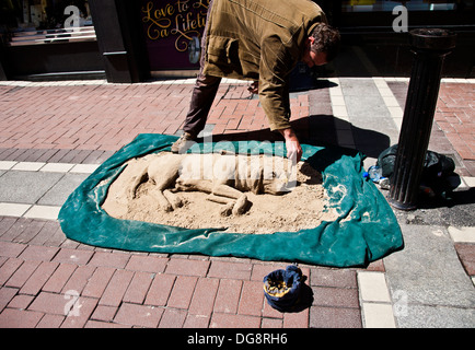 Un uomo che scolpisce l'arte della sabbia in una scultura di sabbia del cane addormentata su Grafton Street a Dublino, Irlanda, Europa, città Street performer arte turismo dell'umorismo Foto Stock