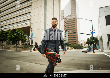 Giovane imprenditore che trasportano skateboard e attraversando via Foto Stock