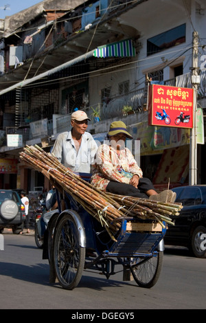 Un uomo è peddling un ciclo con un passeggero e un carico di bambù su una strada di città in Phnom Penh Cambogia. Foto Stock