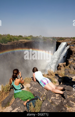 Due donne guardando oltre il bordo a Victoria Falls con arcobaleno, Zambia lato, Livingstone Island, Zambia Africa Foto Stock