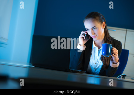 Donna in ambiente office funzionante a una scrivania con un computer portatile ed una tazza di caffè durante la notte mentre utilizzando un iphone Foto Stock