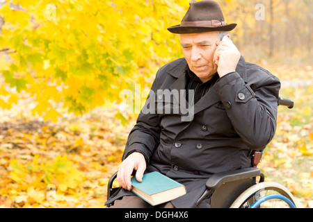 Anziani disabili uomo seduto nella sua sedia a rotelle in un colorato parco autunno utilizzando un telefono cellulare ascoltare attentamente per la conversazione. Foto Stock