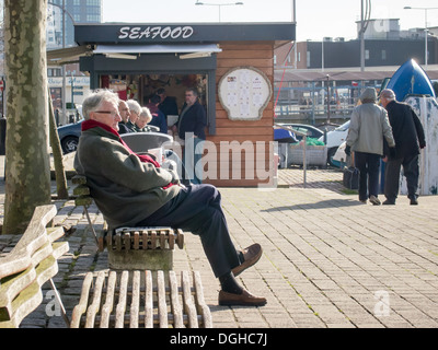 Un uomo che indossa abiti caldi solo seduta su una panchina sul disco di Portsmouth con una pressione di stallo di frutti di mare in background Foto Stock