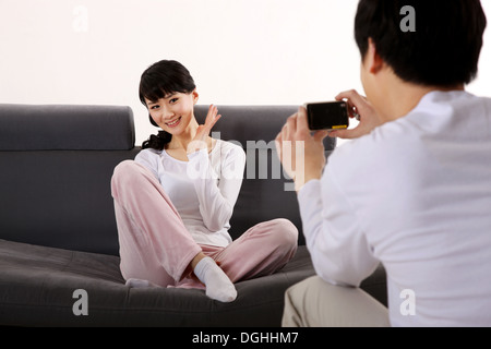 East Asian matura in bianco abiti casual, uomo prendendo foto della donna seduta sul divano Foto Stock