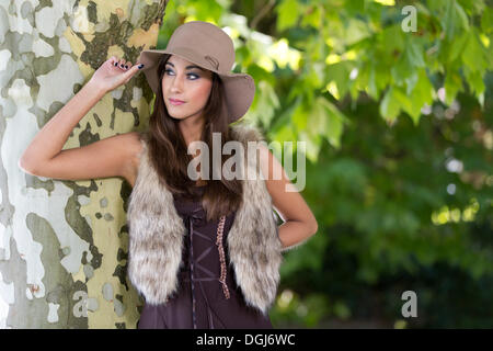 Giovane donna con cappello, fake fur gilet e un abito marrone che pongono in una struttura ad albero Foto Stock