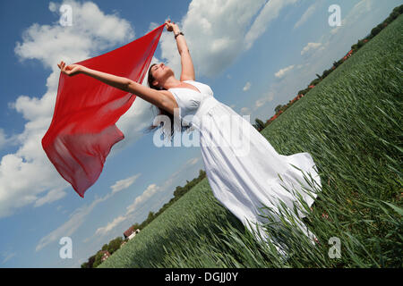Vista obliqua di una giovane donna che indossa un bianco abiti estivi nel verde di un campo di mais mentre si tiene un rosso foulard di seta che fluttua nel Foto Stock