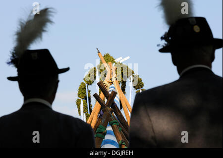 Due uomini che indossano costumi tradizionali a guardare il maypole essendo impostato in Holzhausen, Muensing, Alta Baviera, Baviera Foto Stock