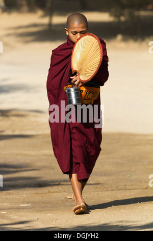 Un monaco novizio con ventilatore, lunchbox e recipiente di raccolta a Bagan in Myanmar. Foto Stock