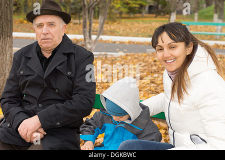 Sorridente cordiale donna seduta su una tavola di legno una panchina nel parco con il suo giovane figlio e padre anziano come loro godere di un tonificante autunno il giorno all'aria fresca. Foto Stock