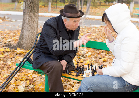 Uomo anziano sostenendo durante una partita a scacchi lo scuotimento di un dito in corrispondenza di una giovane donna come i due si siedono su una tavola di legno una panchina nel parco circondato da caduta foglie di autunno. Foto Stock